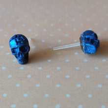 Load image into Gallery viewer, A close of picture of metallic sapphire colored skull earrings displayed on a pale orange background with white polka dots.