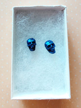 Load image into Gallery viewer, A close of picture of metallic sapphire colored skull earring in a white paper jewelry box.