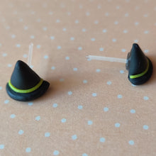 Load image into Gallery viewer, A pair of black witches hats with yellow bands made into flat backed stud earrings. Earrings are displayed on a pale orange background with white polka dots.