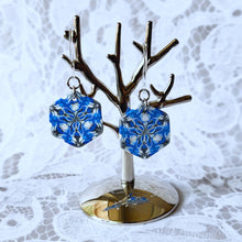 Load image into Gallery viewer, A pair of blue, white, and silver kaleidoscope hexagon earrings hangs from a small polished silver tree stand.