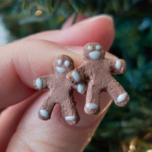 Load image into Gallery viewer, A pair of earrings made to look like gingerbread men. The earrings are held between finger and thumb in front of an artificial Christmas tree.