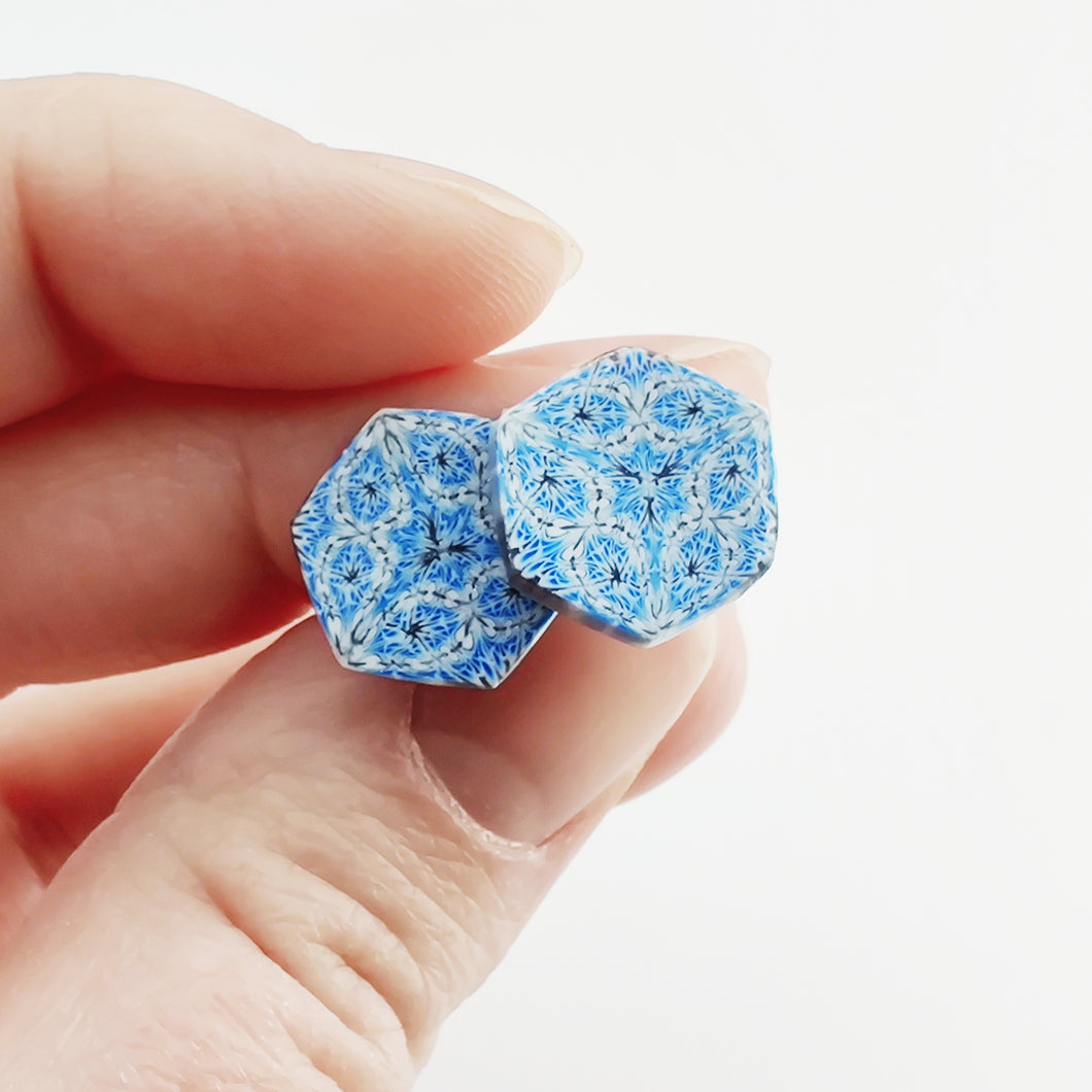 Two hexagon shaped earrings with intricate white, silver and blue patterns held between finger and thumb.